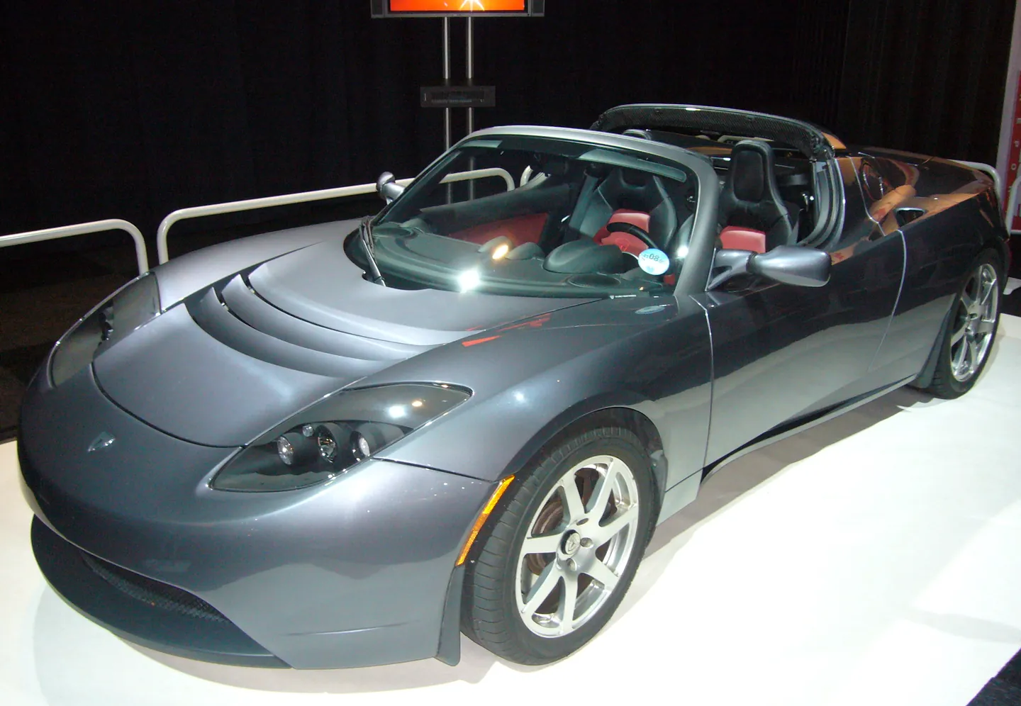 Gray 2008 Tesla Roadster convertible with a red and tan interior on a display stand, seen from the front corner
